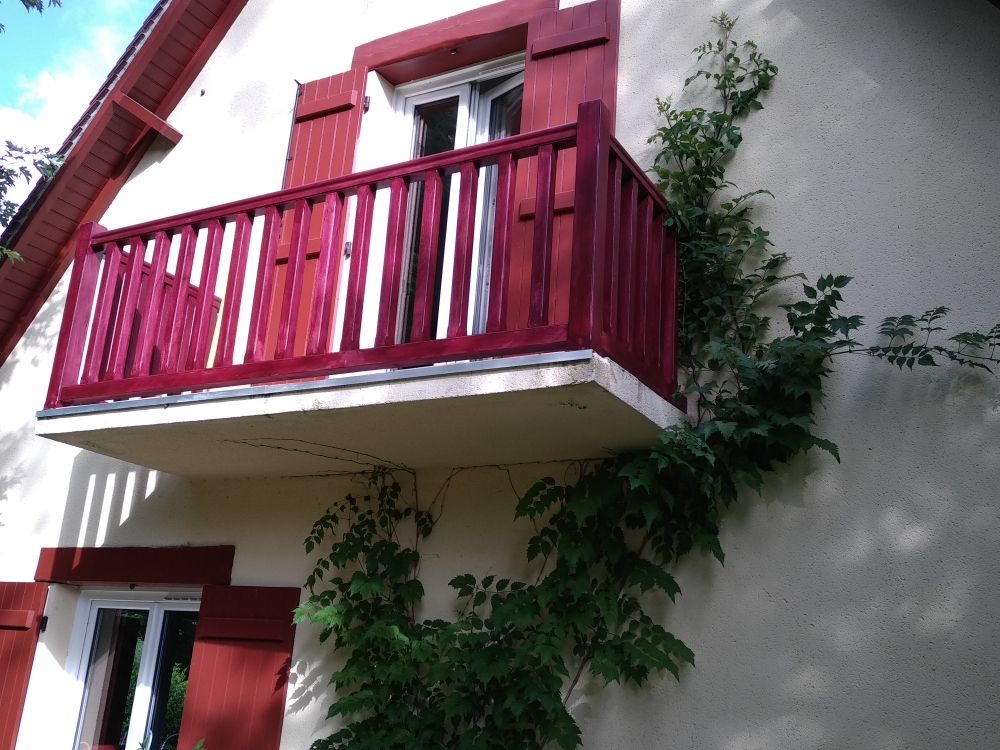 Une façade de maison couleur crème avec un balcon rouge et des volets assortis, et une vigne grimpant le long du mur.