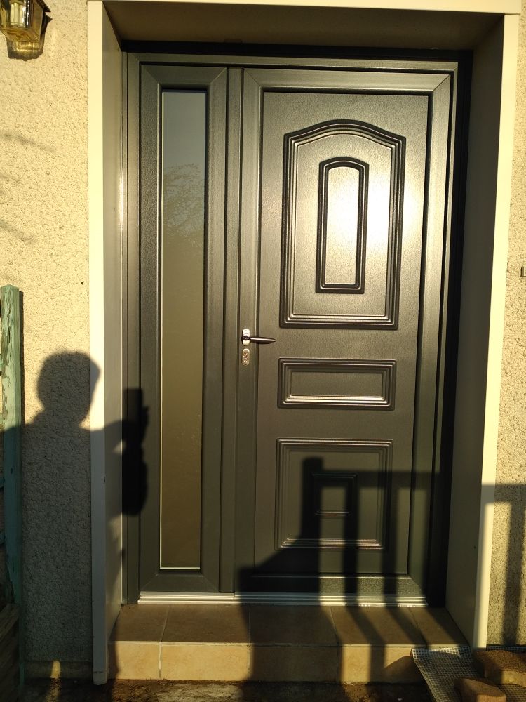Une porte d'entrée gris foncé avec un panneau de fenêtre latéral assorti, encastrée dans un mur extérieur de couleur claire.