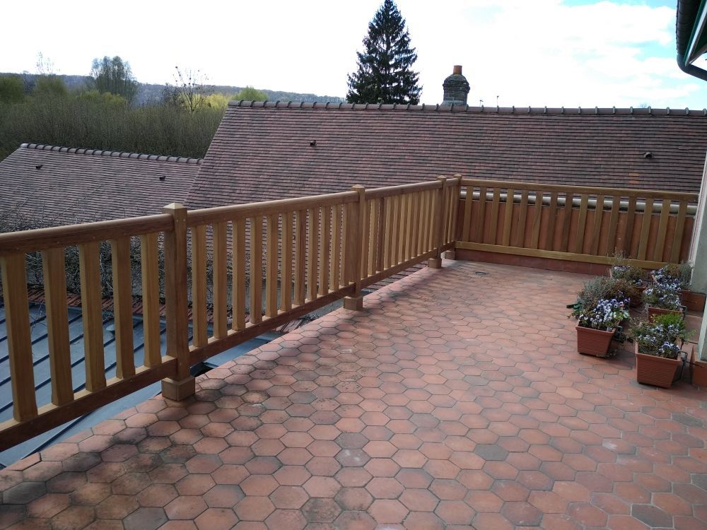 Un balcon extérieur en bois avec un sol en carreaux hexagonaux, donnant sur un toit de tuiles et des arbres.