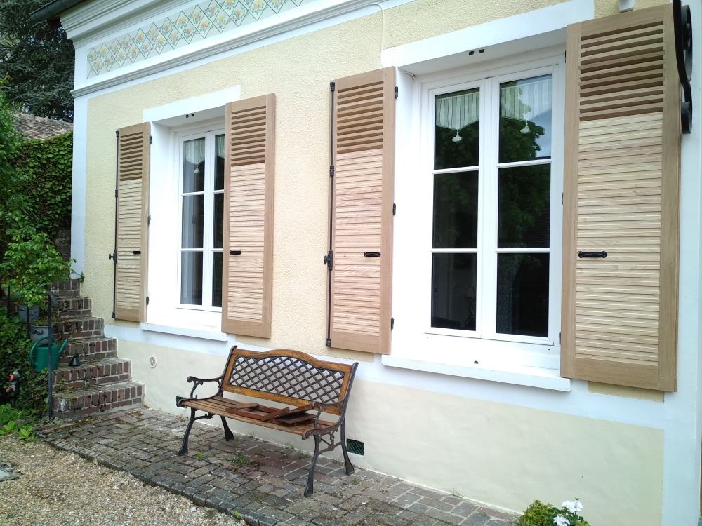 Extérieur d'une maison beige présente deux fenêtres à cadre blanc, des volets persiennes couleur sable et un banc décoratif en métal sur la terrasse.