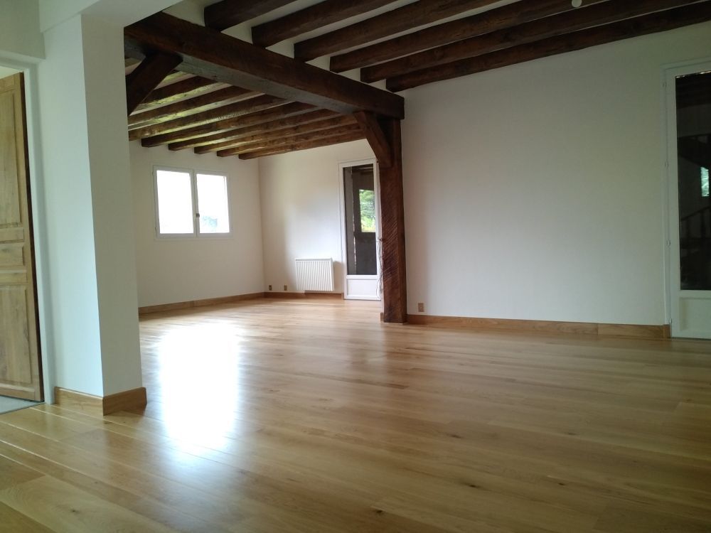 Une pièce vide avec un parquet clair, des murs blancs et un grand plafond aux poutres apparentes en bois sombre.