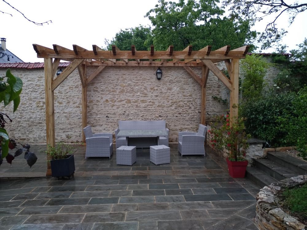 Une pergola en bois abrite un ensemble de patio composé d'un canapé, de deux chaises et de deux poufs, sur une terrasse carrelée en pierre, près d'un mur en pierre.