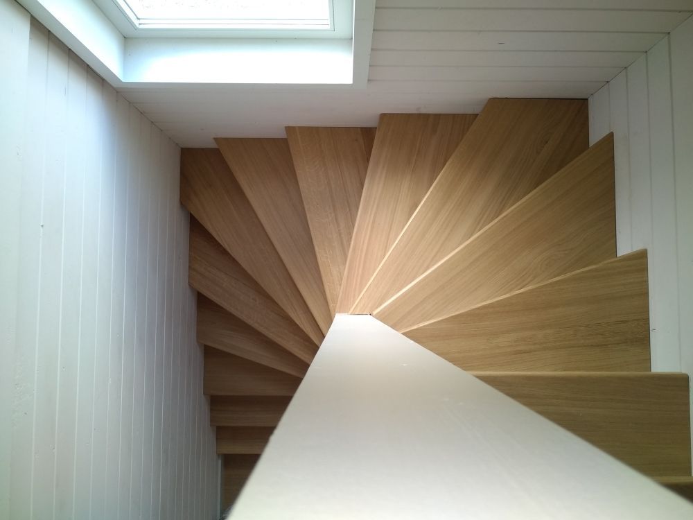 Vue en plongée d'un escalier en colimaçon étroit en bois, aux murs lambrissés blancs et à la rampe blanche.