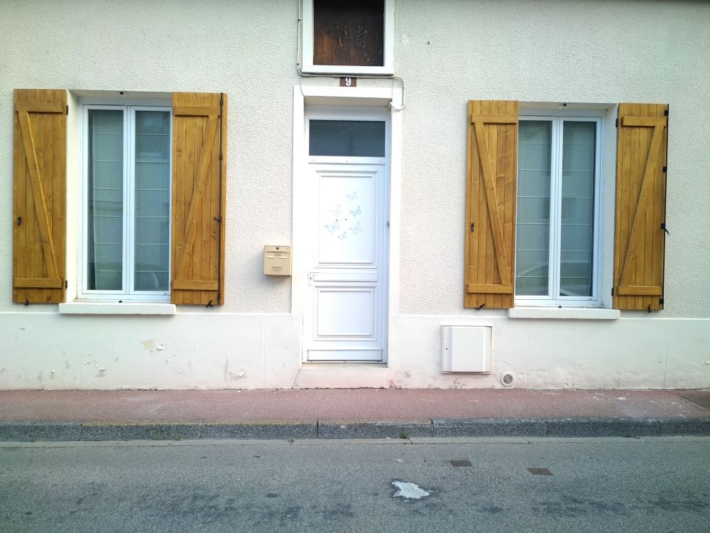 Une façade de maison blanche avec une porte centrale et une fenêtre de chaque côté, agrémentée de volets en bois et d'un petit trottoir.