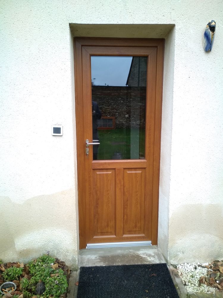 Une porte à cadre en bois brun, avec une vitre en partie supérieure et une base pleine, encastrée dans un mur extérieur en stuc clair.