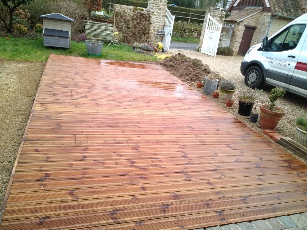 Une terrasse en bois fraîchement installée et encore humide s'étend au-dessus d'une allée, à côté de plantes en pot et d'une camionnette blanche.