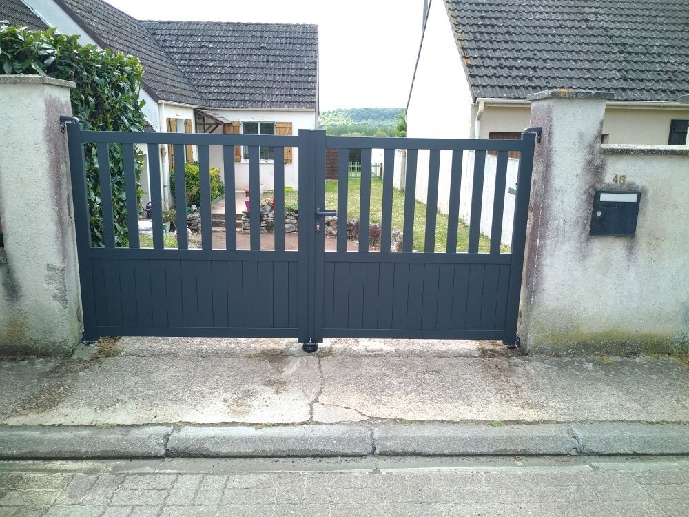 Un portail en métal gris foncé à lattes verticales, situé entre deux piliers de pierre dans une allée résidentielle.