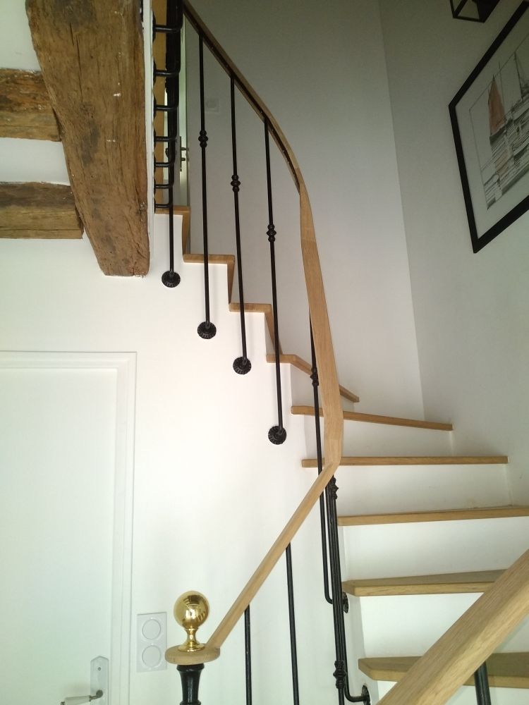 Un escalier en bois avec contremarches blanches, balustres en métal noir, rampe en bois et poteau ornemental doré, dans une pièce lumineuse.