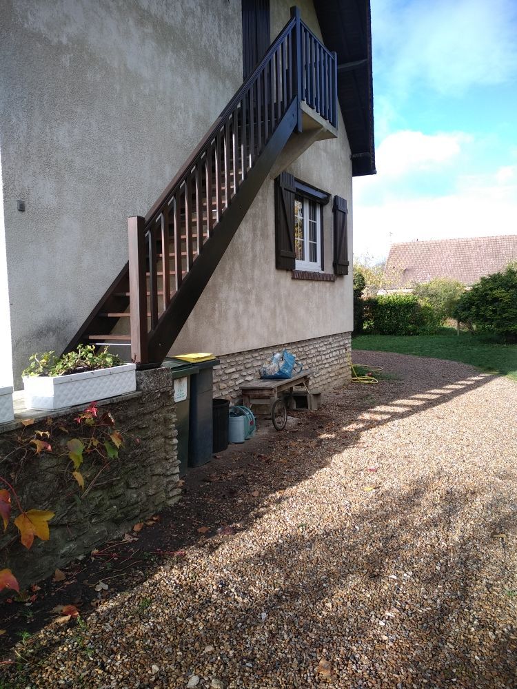 Extérieur d'une maison montrant un escalier extérieur en bois brun foncé menant à un étage supérieur, à côté d'une allée de gravier.