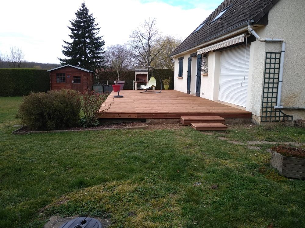 Une terrasse en bois s'étend sur le côté d'une maison jusqu'à une cour arrière gazonnée, avec un abri de jardin et des arbres en arrière-plan.