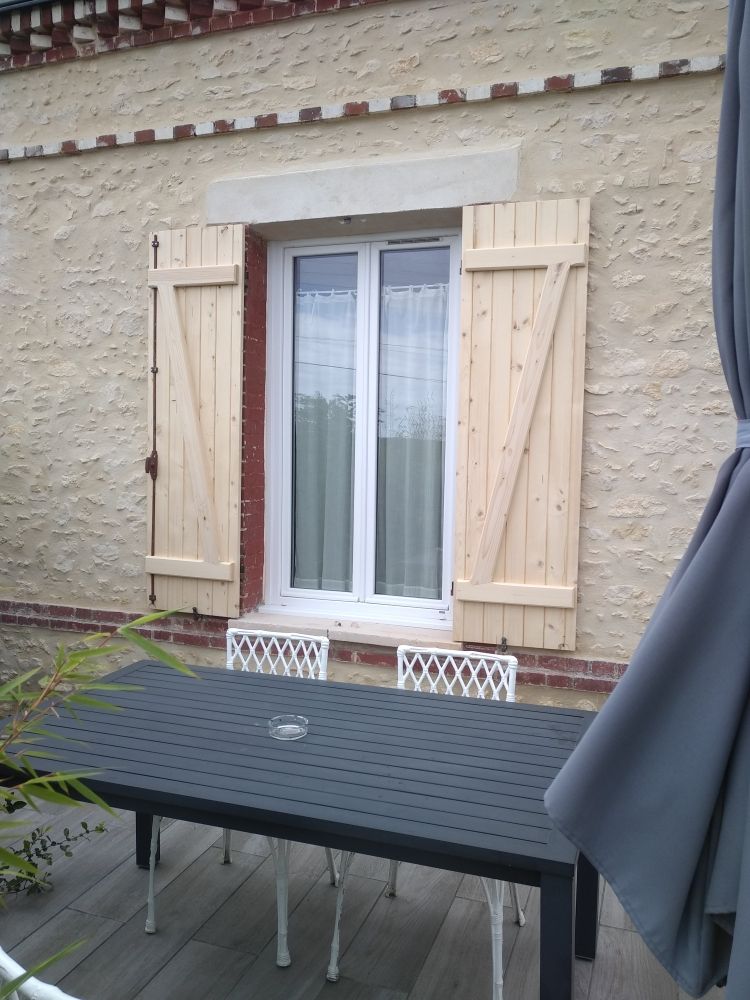Une table et des chaises de patio modernes sont installées sur une terrasse devant le mur d'une maison percée d'une fenêtre blanche et de volets en bois clair.