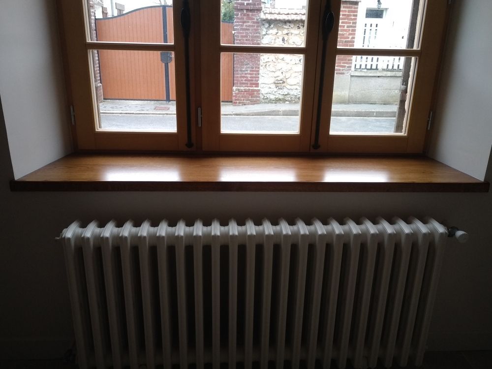 Un appui de fenêtre en bois au-dessus d'un radiateur blanc nervuré, devant une fenêtre donnant sur la rue.