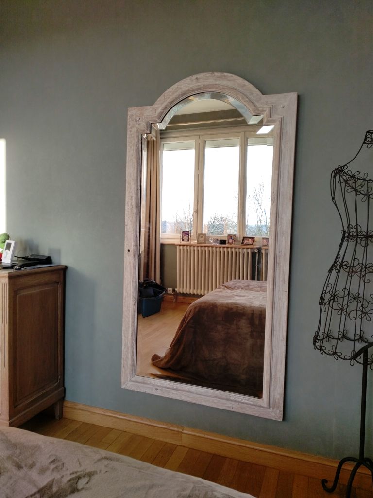 Un grand miroir arqué, au cadre en bois blanc vieilli, est accroché à un mur gris au-dessus d'un parquet clair.