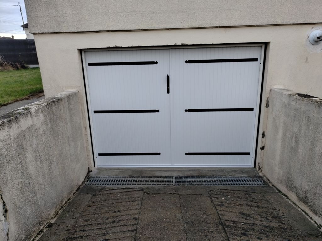 Une porte de garage double blanche ornée de barres horizontales décoratives noires, encastrée dans un mur beige au-dessus d'une allée en béton en pente.