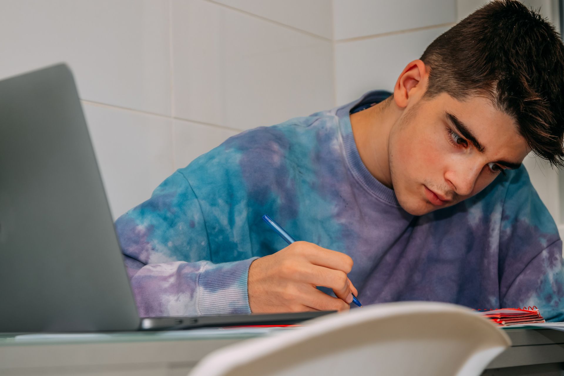 Adolescente con suéter teñido anudado concentrado escribiendo en un escritorio, con una computadora portátil cerca.