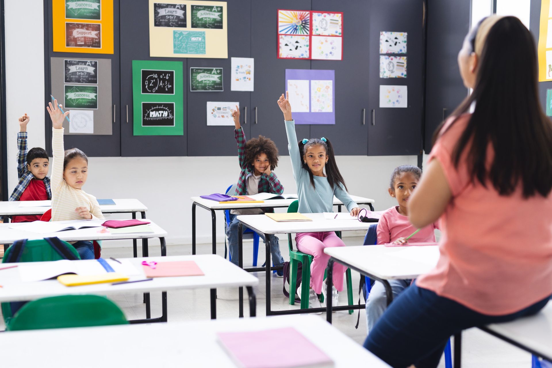Niños levantando las manos en un aula, el profesor al frente.
