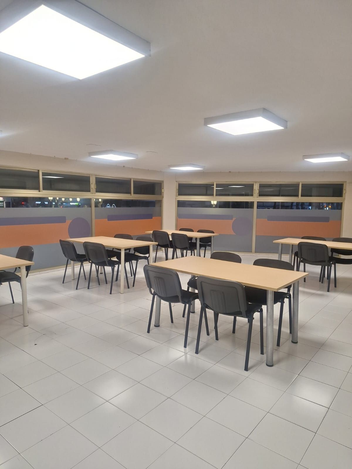Una cafetería vacía con mesas y sillas. Techo y suelo blancos, ventanas con detalles en naranja y morado.