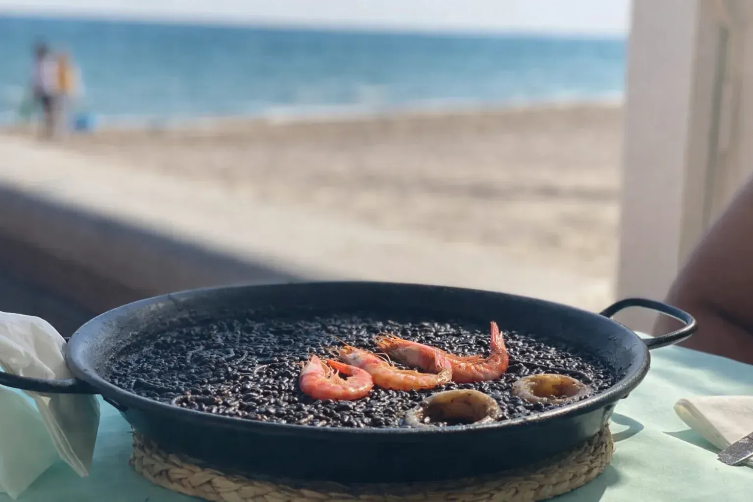 Paella negra con camarones en sartén sobre una mesa, playa y océano de fondo.