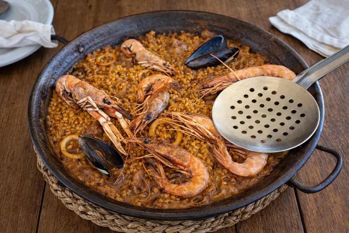 Paella en sartén negra con camarones, mejillones y calamares sobre una tabla de madera, con una espumadera.