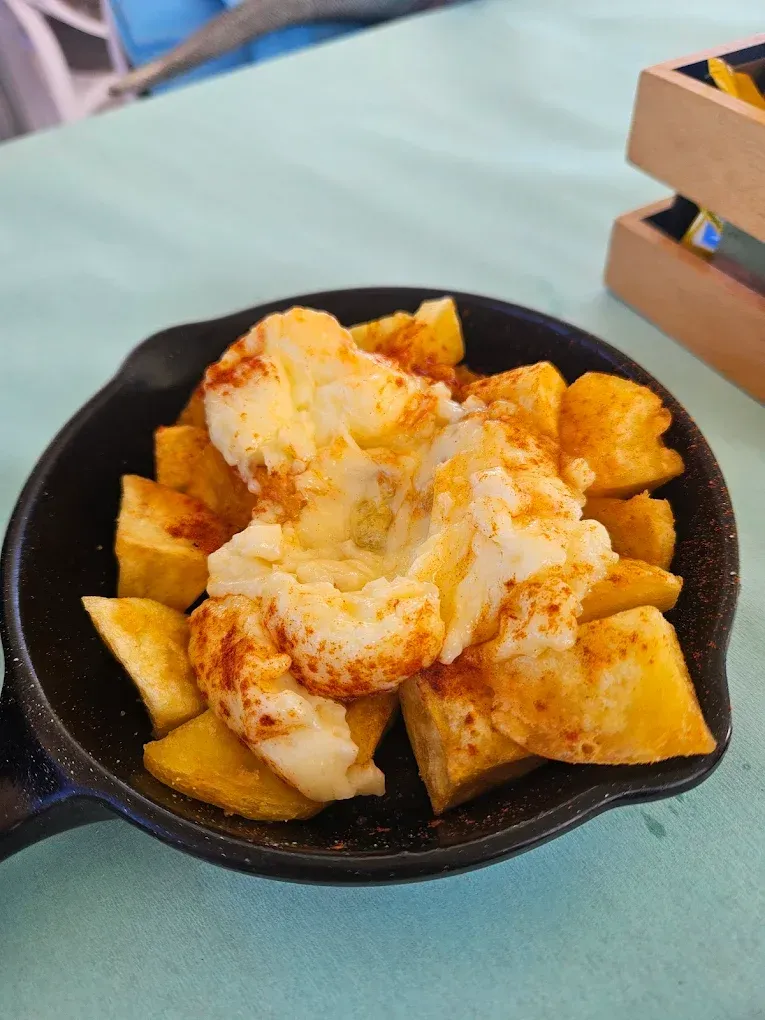 Patatas bravas con alioli en sartén negra, espolvoreadas con pimentón, sobre un mantel verde azulado.