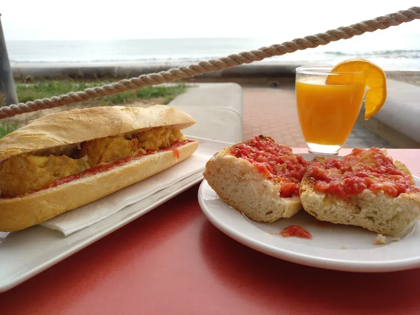 Sándwich, tostada de tomate y zumo de naranja sobre una mesa roja, con la playa al fondo.