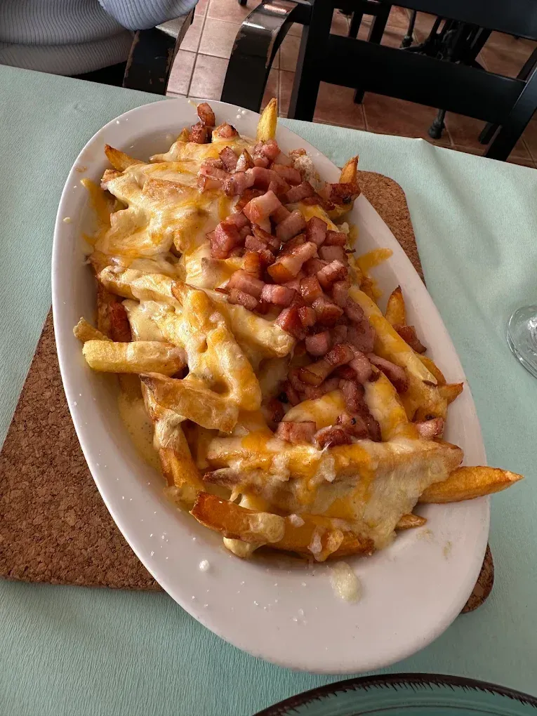 Papas fritas cubiertas con queso derretido y trocitos de tocino en un plato blanco ovalado sobre un posavasos.