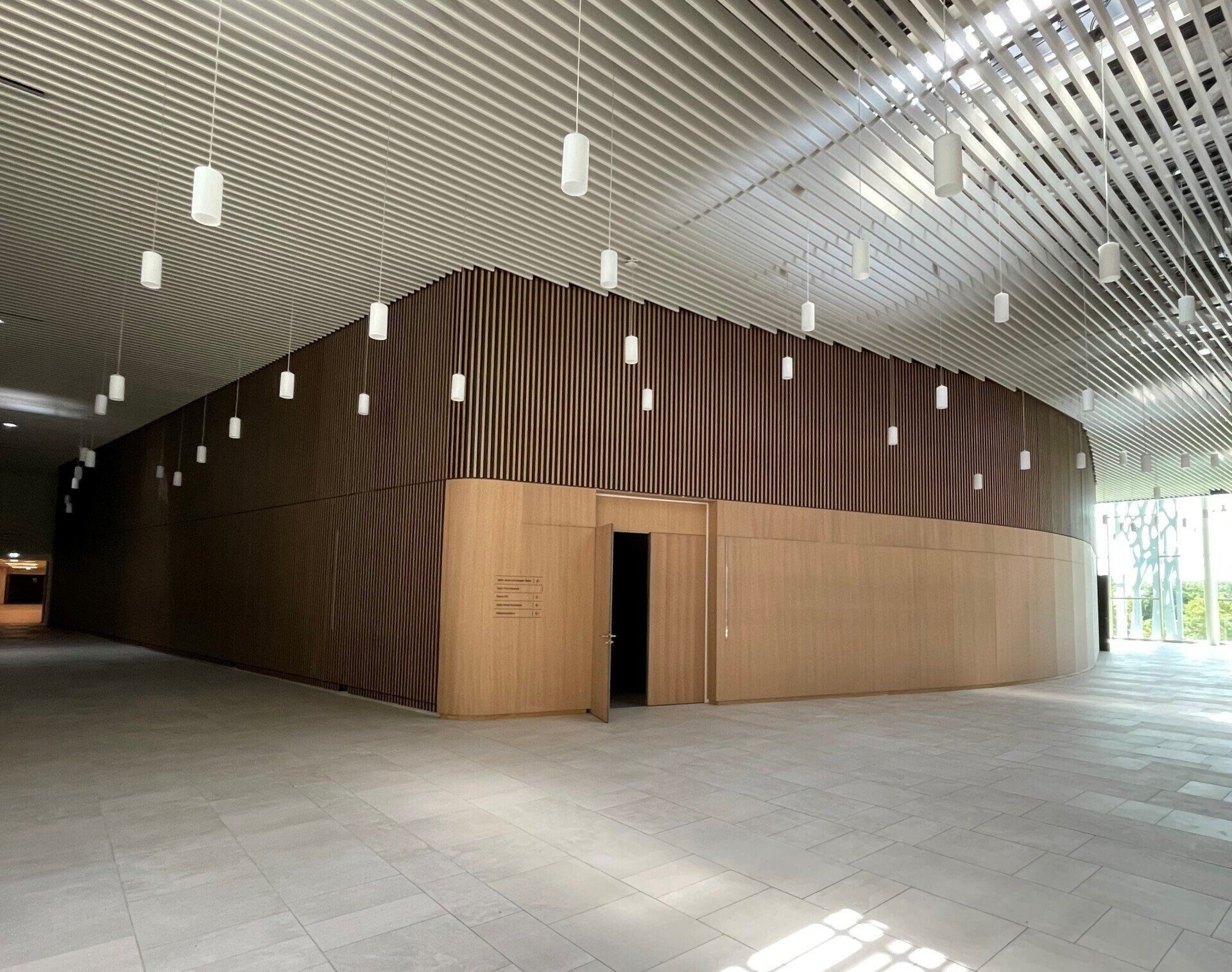 Hall d'entrée avec une structure incurvée en panneaux de bois, un plafond à motifs, des luminaires suspendus et une entrée vitrée.