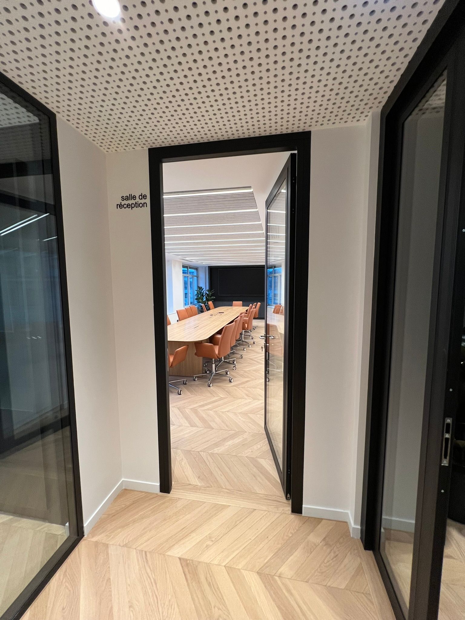 Couloir de bureau avec portes vitrées ouvertes donnant sur une salle de conférence et une longue table.