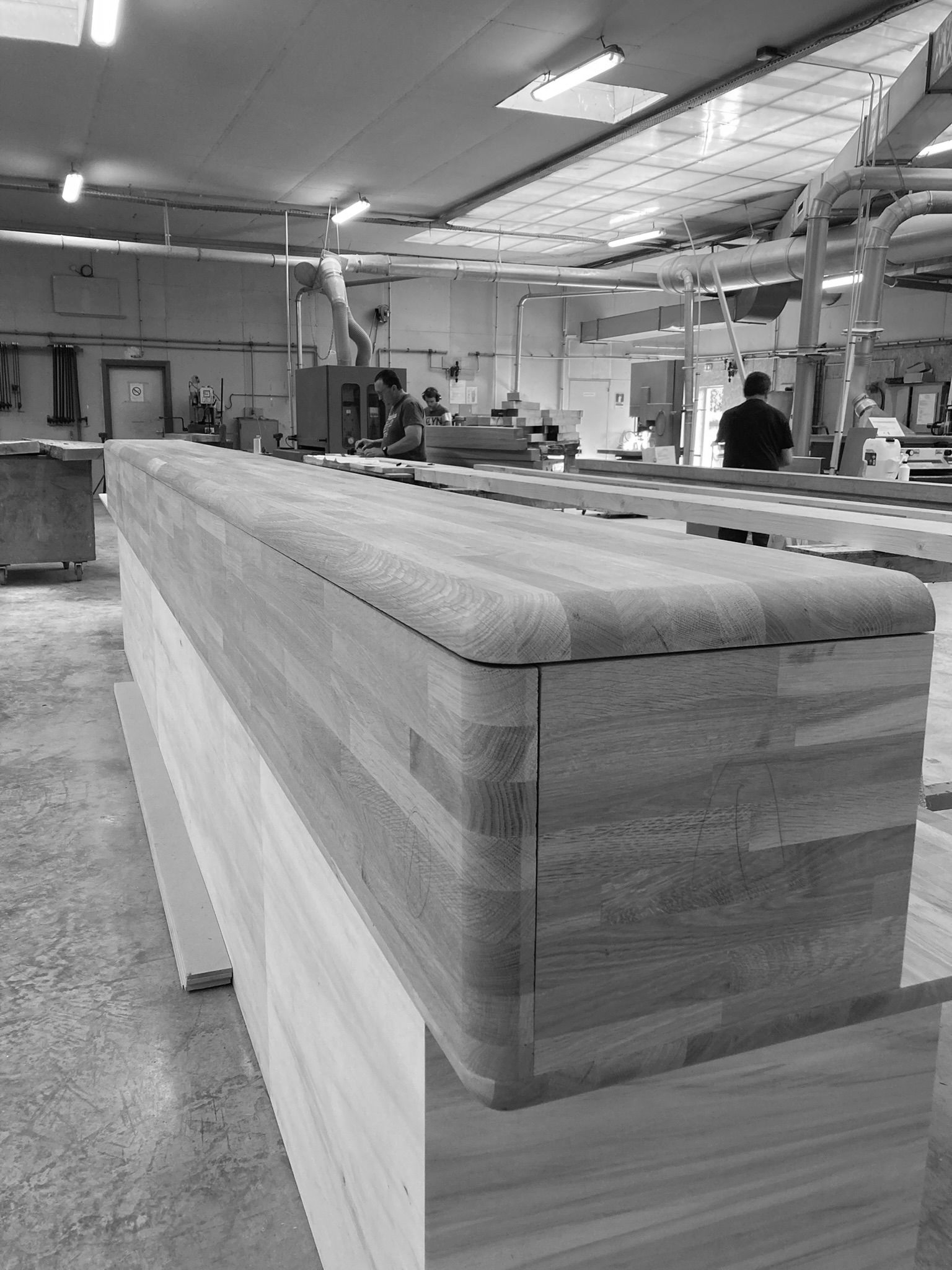 Établi en bois dans un atelier de menuiserie, avec des ouvriers et des outils en arrière-plan.