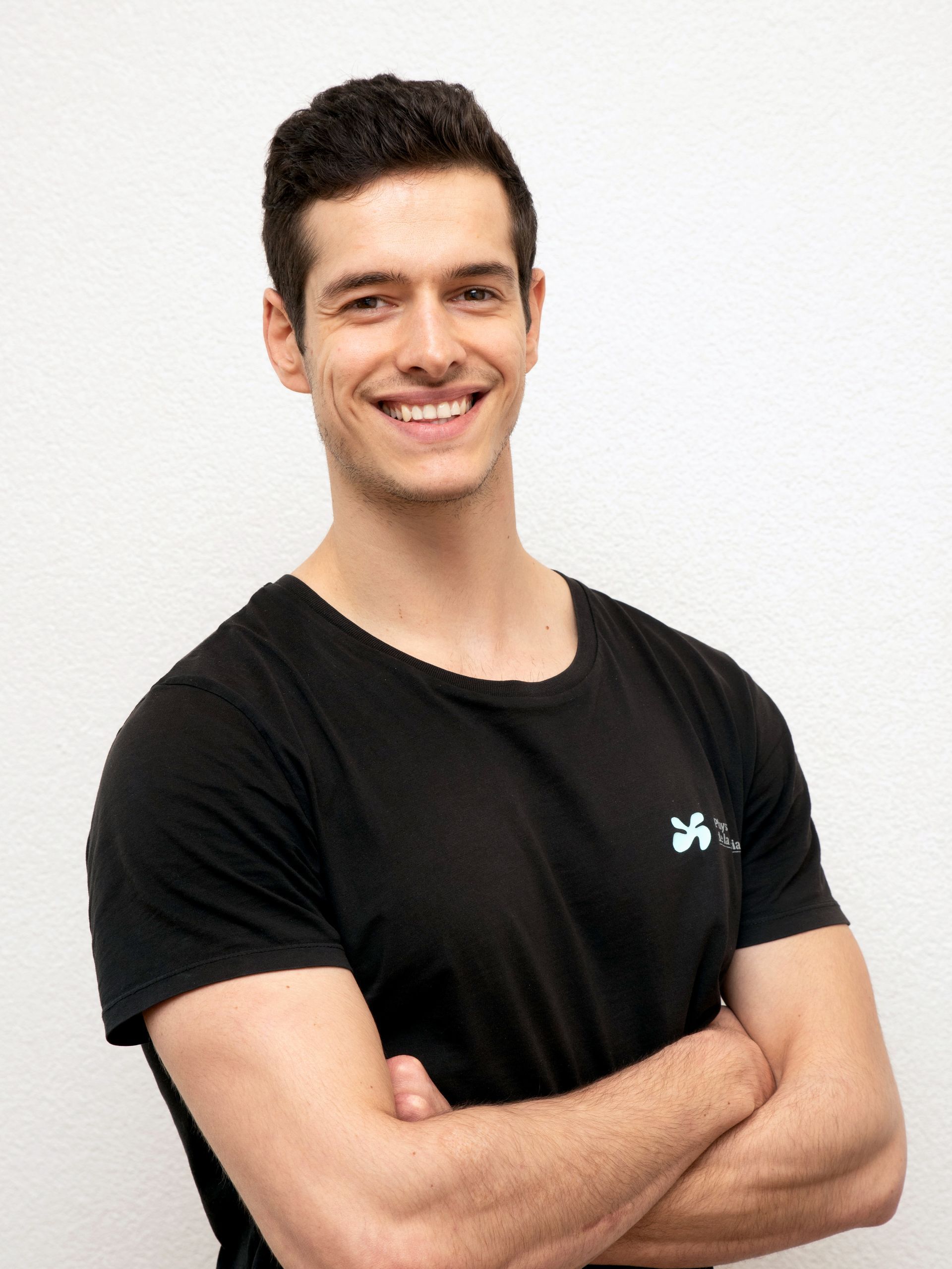 Un homme, les bras croisés, souriant, vêtu d'un t-shirt noir, se tient devant un mur blanc.