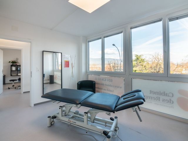 Salle d'examen médical avec lit réglable, miroir et grandes fenêtres donnant sur l'extérieur.