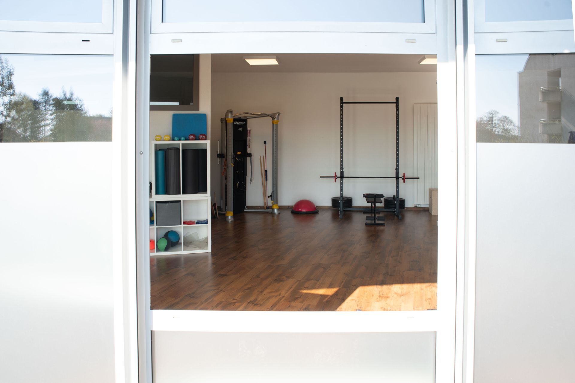 Vue par une fenêtre sur une salle de sport avec des équipements de musculation : haltères, rack, tapis, rouleaux de massage.