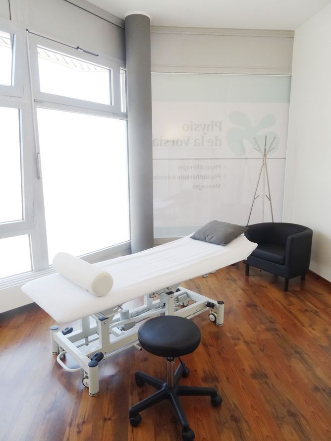 Salle de soins avec table de massage, tabouret, chaise et grande fenêtre.
