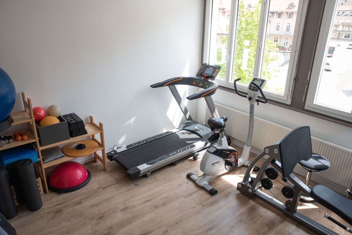 Salle de sport avec tapis roulant, vélo d'appartement et haltères près des fenêtres.