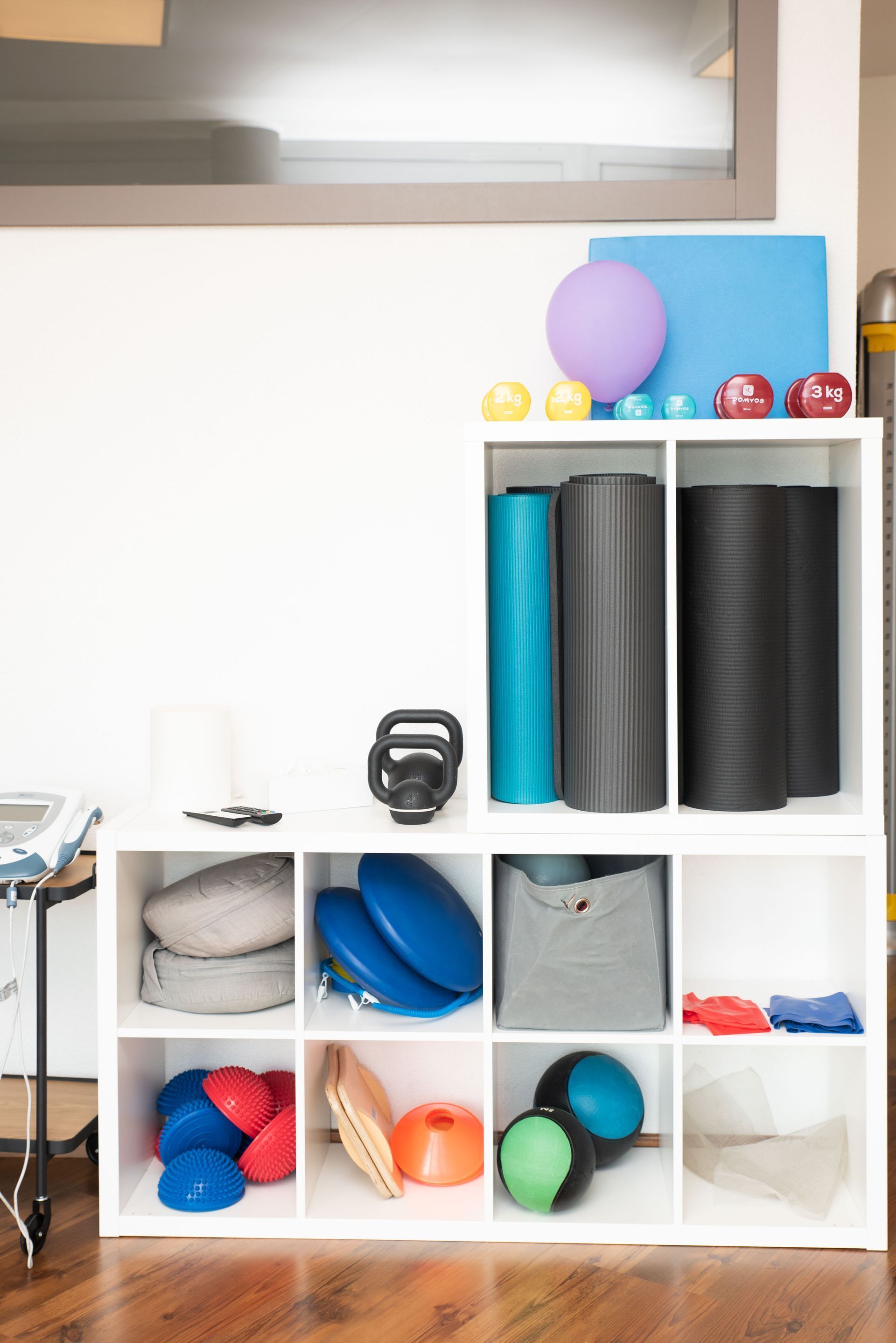 Dans une pièce avec fenêtre, une étagère blanche contient du matériel de fitness : ballons, rouleaux, haltères.