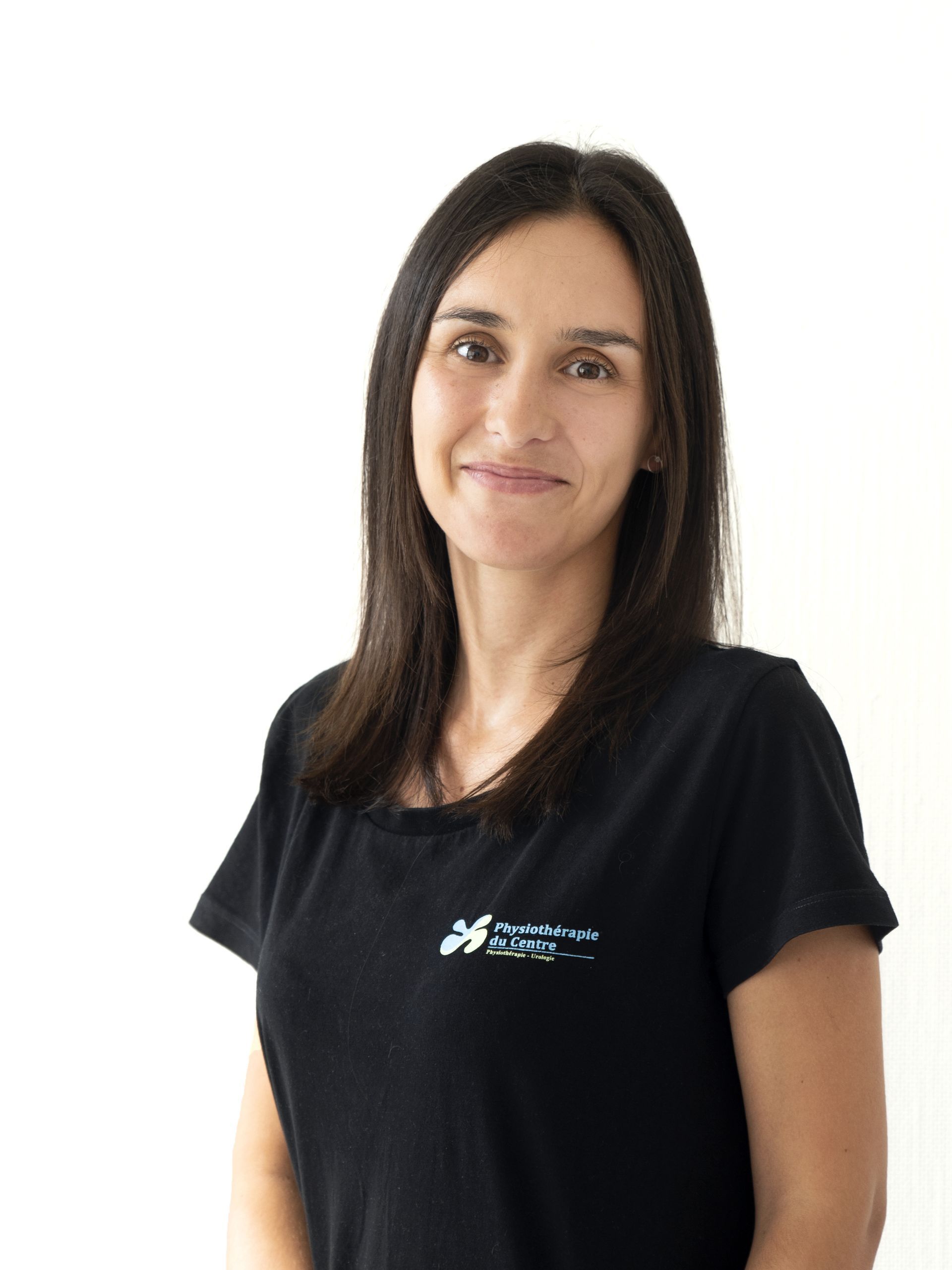 Femme aux cheveux foncés, vêtue d'un t-shirt noir, souriante, face à la caméra sur fond blanc.