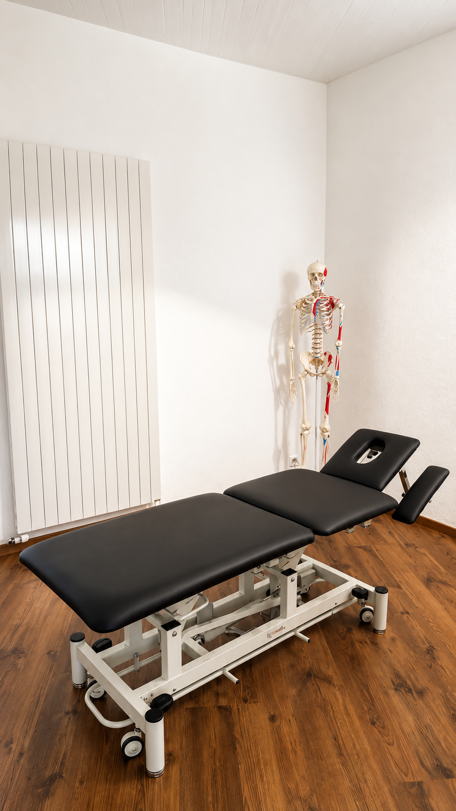 Table de massage noire réglable dans une salle de soins lumineuse avec un modèle d'anatomie squelettique.