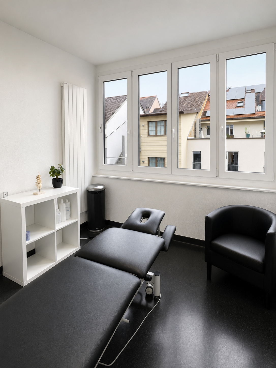 Salle de massage minimaliste avec tables noires, chaise noire, étagère blanche et grandes fenêtres donnant sur les bâtiments