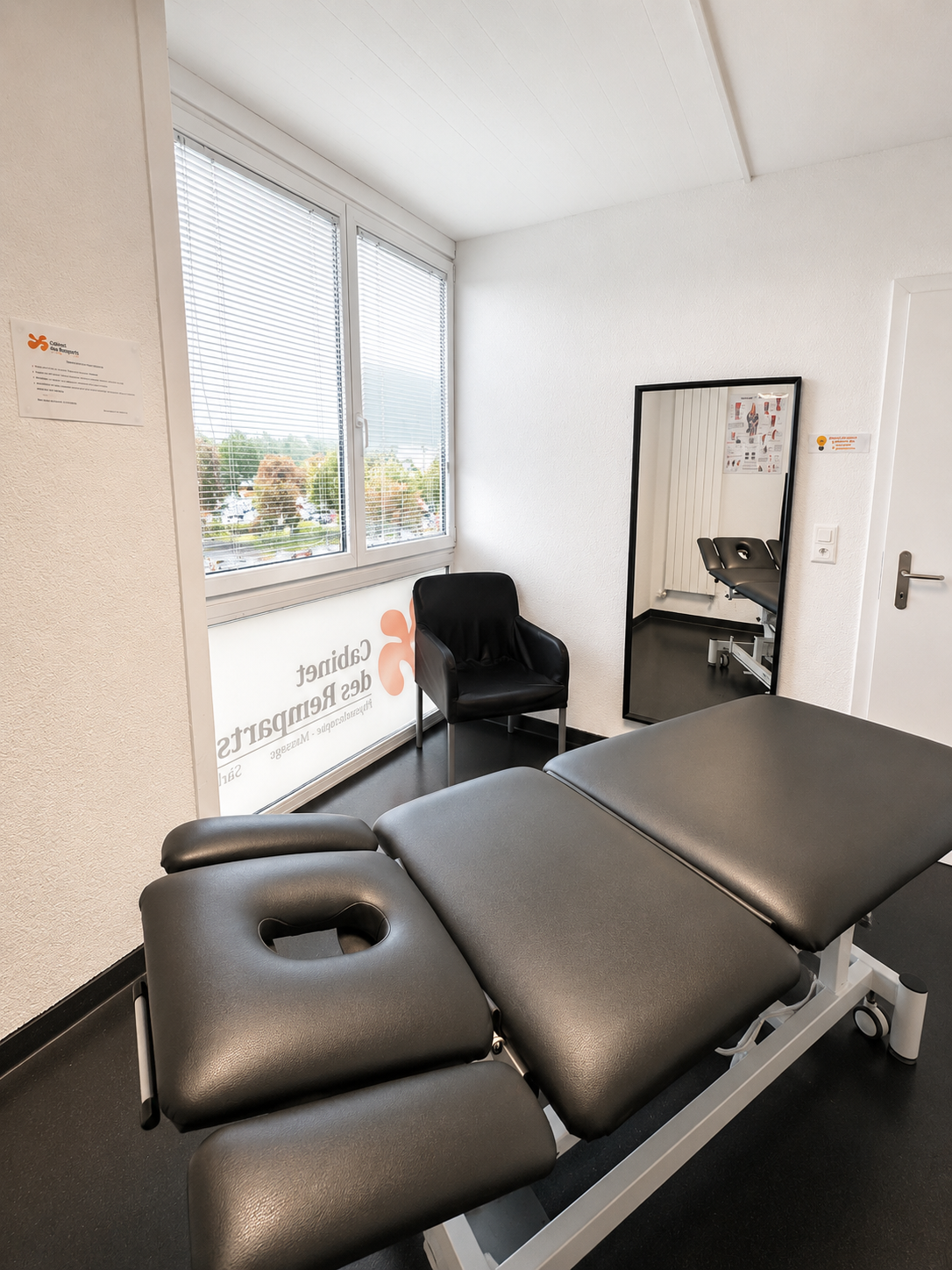 Table de massage noire dans une petite salle de clinique avec une chaise, une fenêtre et un miroir mural.