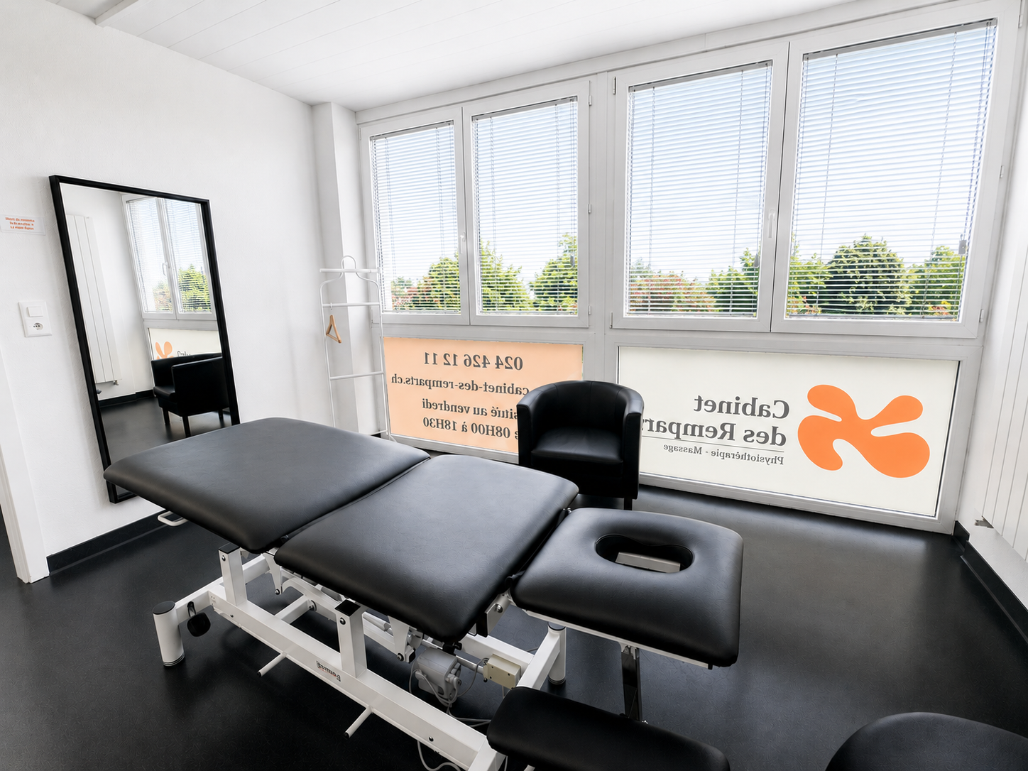 Table de soins dans une salle de clinique lumineuse avec miroir, fenêtres et logo orange sur le mur.