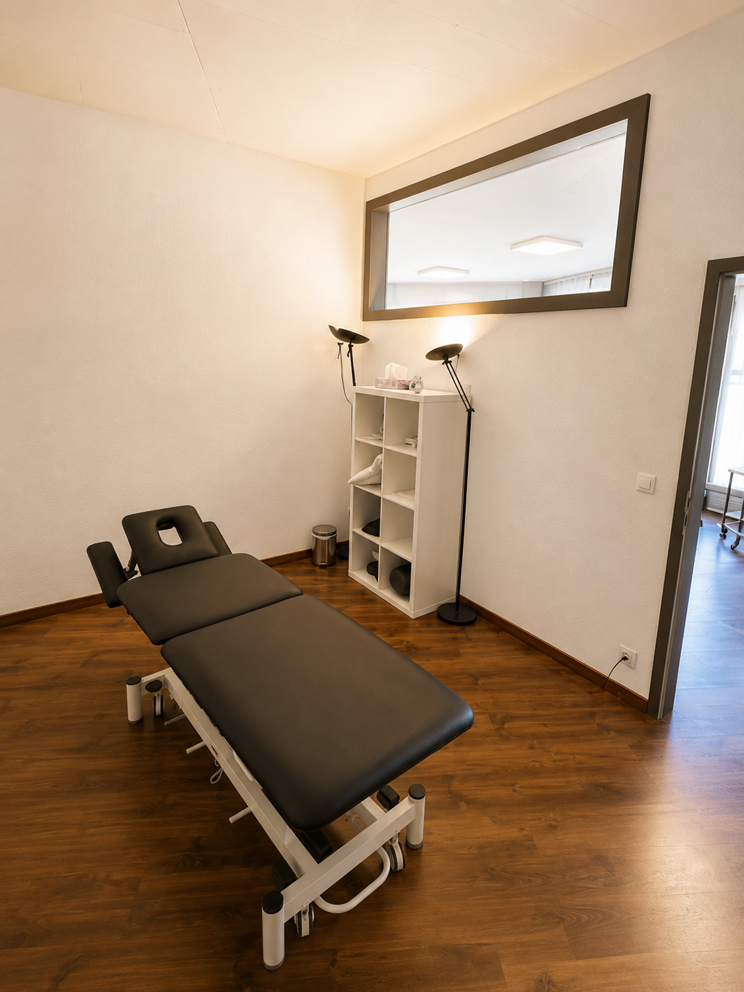Salle de soins avec table de massage noire, étagères blanches et miroir mural sur parquet.