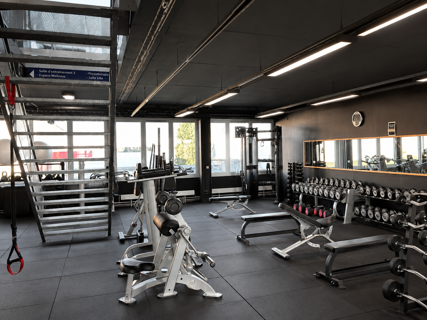 Intérieur de salle de sport moderne avec machines de musculation, bancs, haltères et grandes fenêtres sous un éclairage industriel