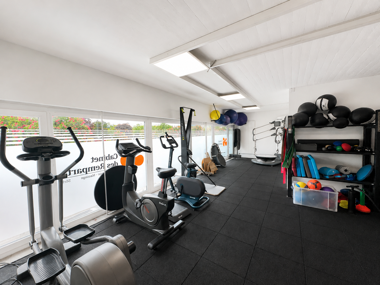 Salle de sport avec vélos elliptiques et rameurs sur un sol en tapis noir, étagères d'équipement le long du mur de droite.