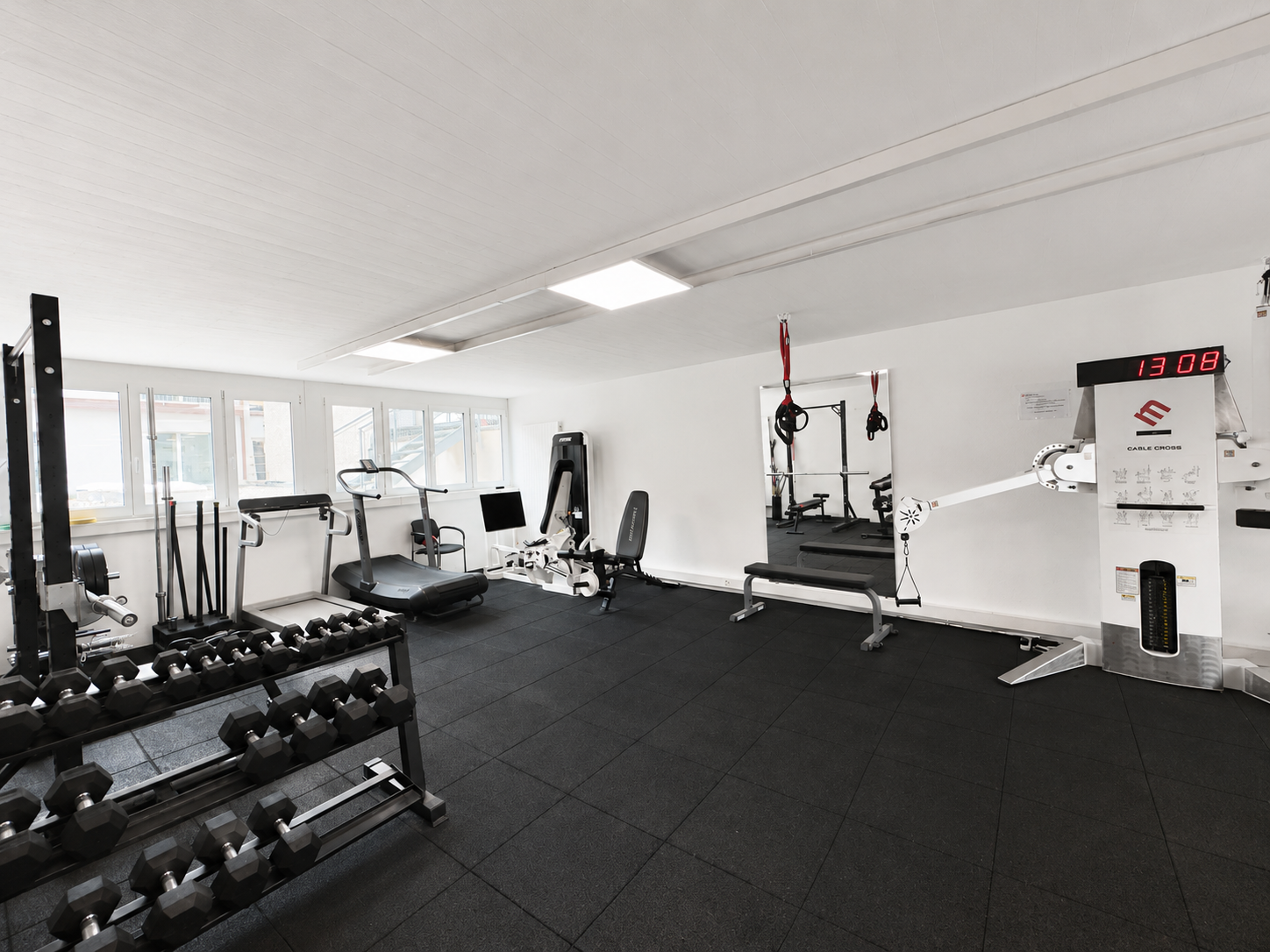 Salle de sport lumineuse avec sol noir, supports à haltères, machines à câbles et équipements de cardio près de grandes fenêtres
