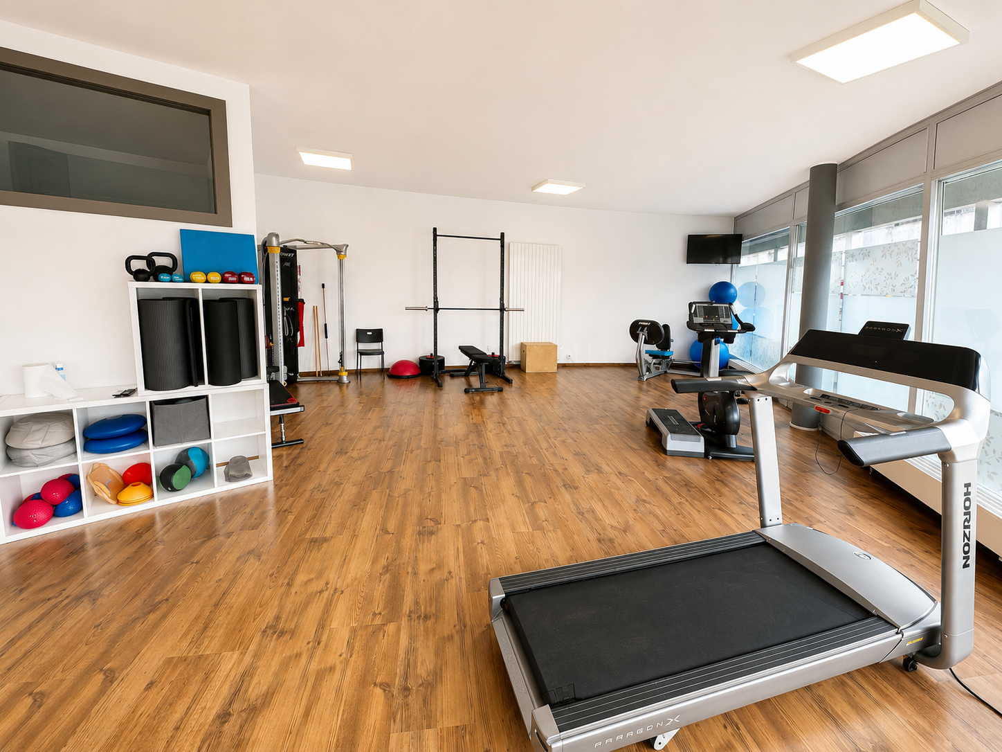 Salle de sport spacieuse avec tapis de course, appareils de musculation et parquet sous de grandes fenêtres.