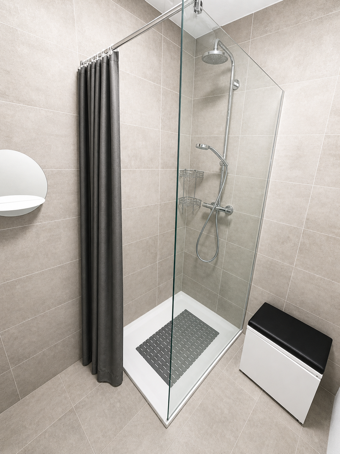 Petite cabine de douche carrelée beige avec porte en verre transparent, rideau gris et poubelle blanche murale dans une salle de bain