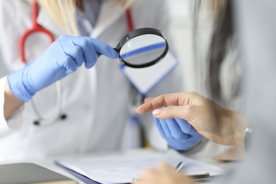 Ein Arzt untersucht die Hand eines Patienten mit einer Lupe. Blaue Handschuhe, weißer Kittel, Stethoskop.