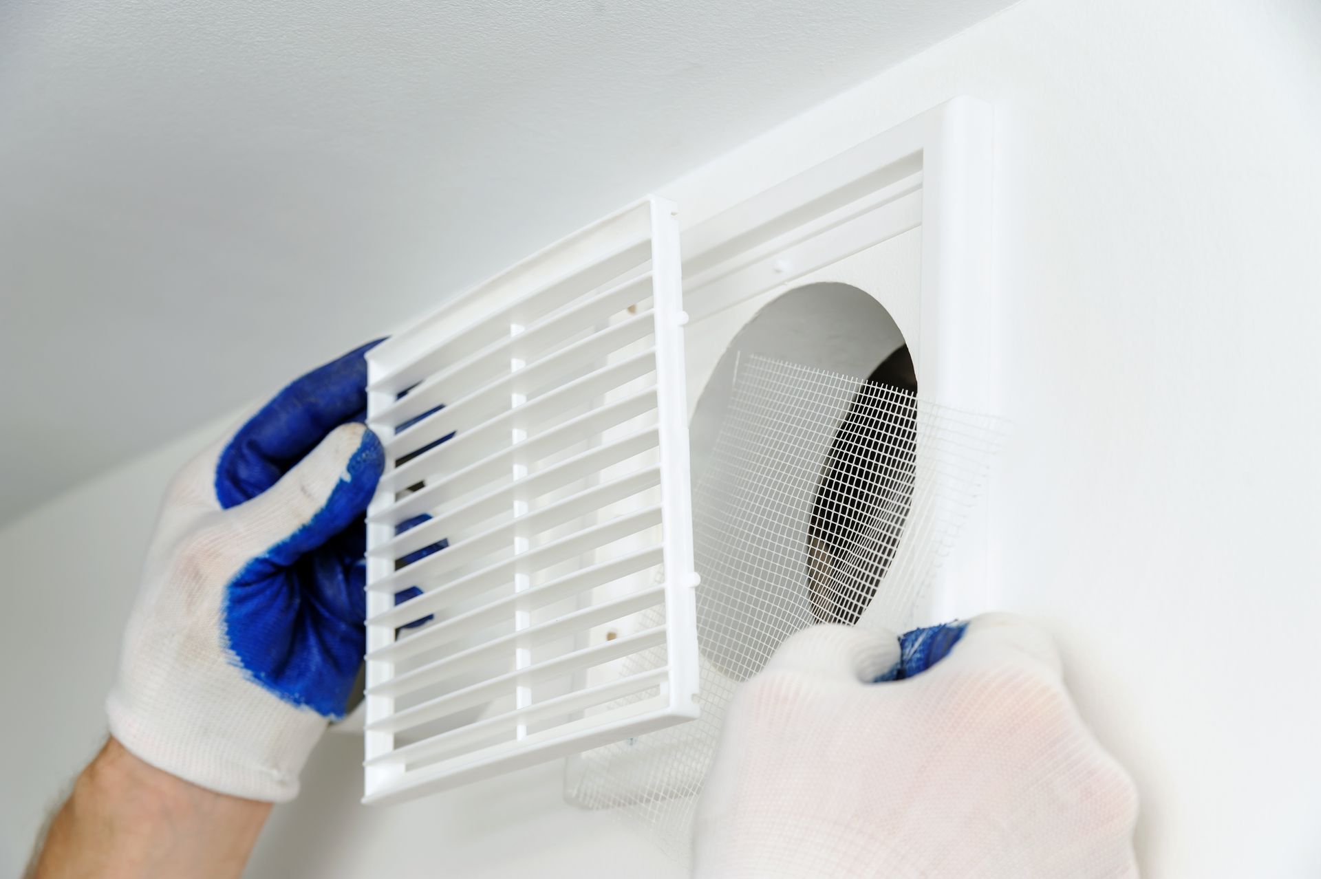 Des mains gantées de bleu nettoient un filtre blanc de grille d'aération au plafond.