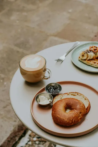 Bagel y café sobre una mesa, con un plato de waffles, en un entorno al aire libre.
