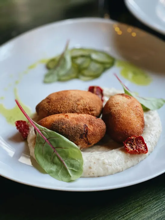Croquetas fritas sobre una cama de puré blanco, con pepinillos, verduras y tomates secos en un plato blanco.